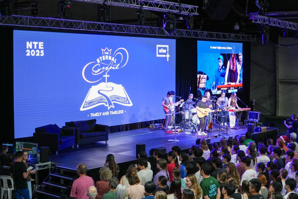 A band playing on a conference main stage in front of a wide LED wall carrying the event's branding, with a second screen beside it relaying a close-up of the singers to a standing crowd.