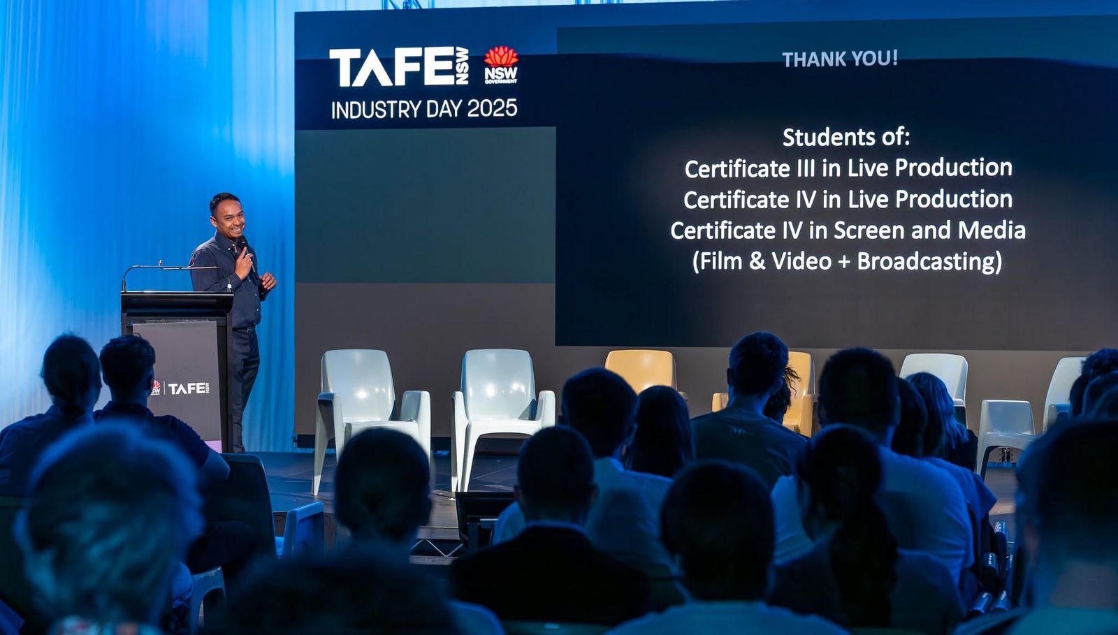 A presenter at a lectern beside a wide LED wall carrying the event's slides at a TAFE industry day, rows of students watching under blue stage lighting.