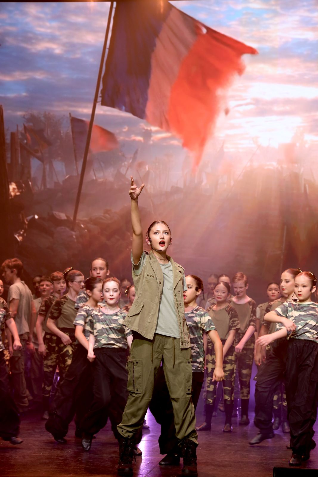 A young performer with one arm raised leads a costumed ensemble, the LED wall behind them showing a sunrise battlefield backdrop in place of built scenery.