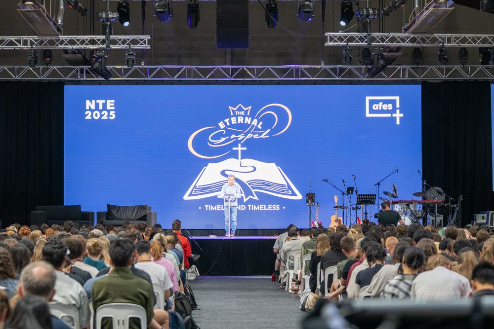 A speaker at a lectern on a conference stage in front of a wide LED wall carrying the event's branding, with several hundred people seated in front of it.
